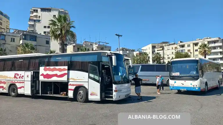 A large high-capacity intercity coach parked at Durrës terminal bound for Tirana, showing all passenger doors and the side luggage hold open. A group of bus drivers stands nearby in conversation.